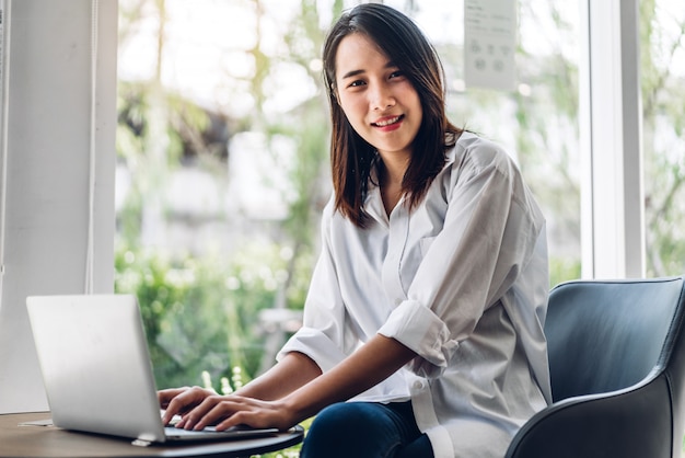 Portret van lachende gelukkig mooie aziatische vrouw ontspannen met behulp van laptopcomputer zittend op de bank. jonge hipster meisje freelancer werken en denken met nieuwe ideeën in café