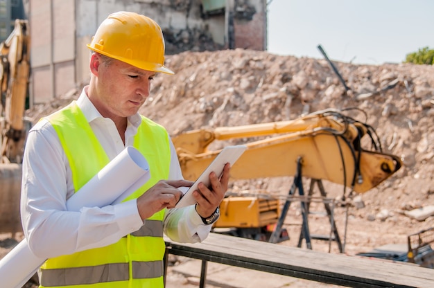 Portret van lachende jonge architect of knappe ingenieur bedrijf tablet en opgerolde blauwdrukken met bouwconstructie achtergrond.