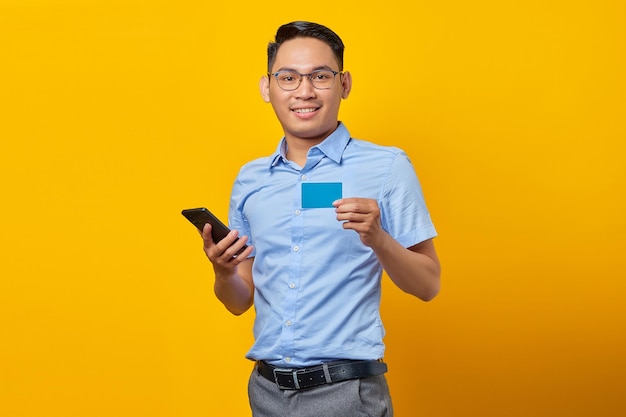 Portret van lachende jonge Aziatische man Aziatisch in glazen met mobiele telefoon en creditcard geïsoleerd op gele achtergrond zakenman en ondernemer concept