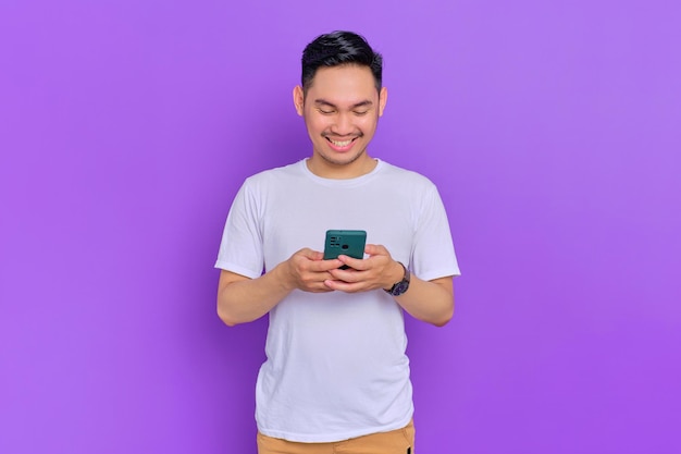 Portret van lachende jonge Aziatische man in witte t-shirt met behulp van smartphone voor messaging met vrienden geïsoleerd op paarse achtergrond