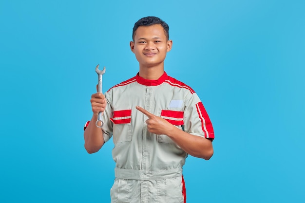 Portret van lachende jonge Aziatische monteur wijzende moersleutel met vinger geïsoleerd op blauwe achtergrond