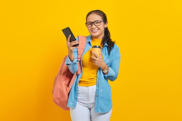Portret van lachende jonge Aziatische studente in denim kleding met rugzak met kopje koffie en het gebruik van smartphone geïsoleerd op gele achtergrond