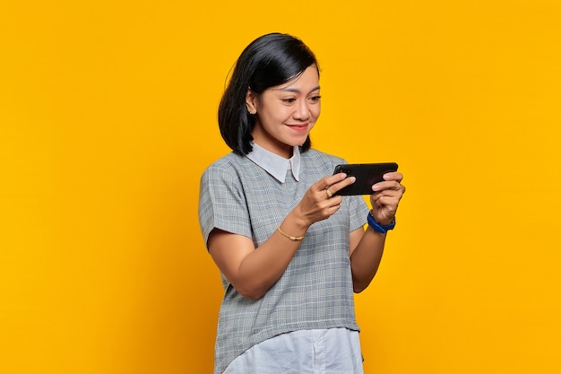 Portret van lachende jonge Aziatische vrouw die videogame speelt op mobiele telefoon over gele achtergrond