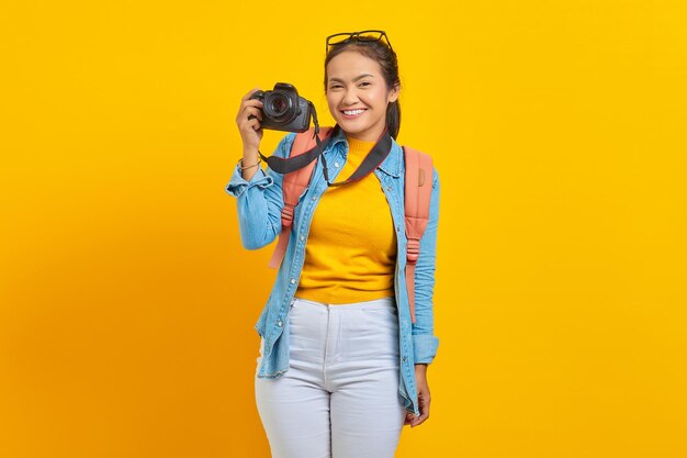 Portret van lachende jonge Aziatische vrouw in denim kleding met rugzak en het tonen van professionele camera geïsoleerd op gele achtergrond