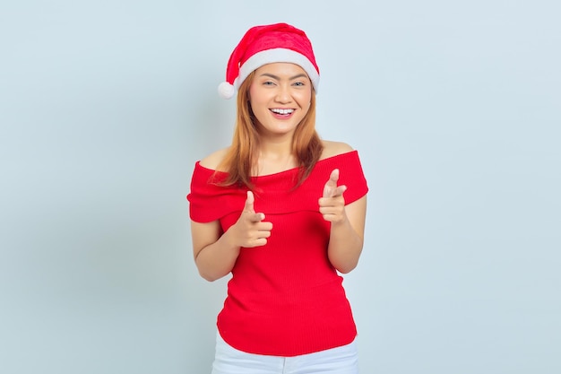 Portret van lachende jonge Aziatische vrouw met rode jurk en kerstmuts wijzende vinger naar camera geïsoleerd op een witte achtergrond