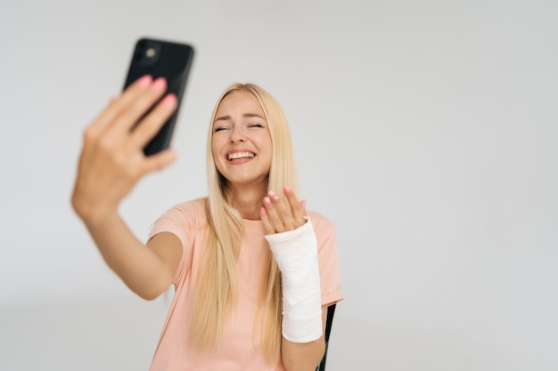 Portret van lachende jonge blonde vrouw met gebroken arm gewikkeld in gipsverband pratende smartphone die videogesprek voert en selfie-foto maakt