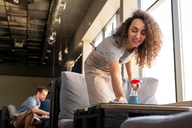 Portret van lachende jonge SMM manager met krullend haar zittend op een comfortabele bank en werken met laptop in café