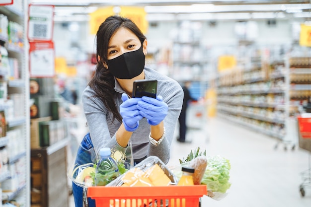 Portret van lachende jonge vrouw in beschermend masker en handschoenen smartphone in handen houden tijdens het winkelen in de supermarkt.