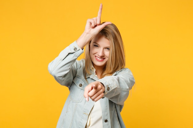 Portret van lachende jonge vrouw in denim casual kleding met verliezer gebaar, wijzende wijsvinger op camera geïsoleerd op geeloranje muur achtergrond. Mensen levensstijl concept. Bespotten kopie ruimte.