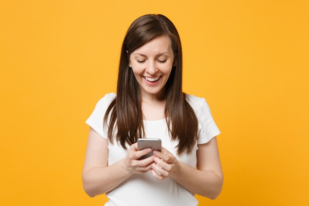 Portret van lachende jonge vrouw in witte casual kleding met behulp van mobiele telefoon, sms-bericht typen geïsoleerd op fel geel oranje muur achtergrond in studio. Mensen levensstijl concept. Bespotten kopie ruimte.