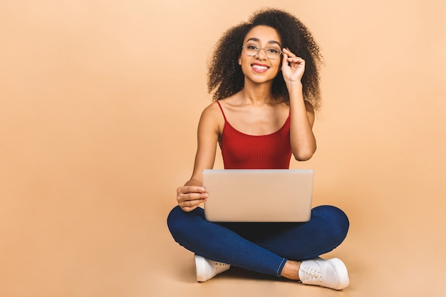 Portret van lachende jonge vrouw met behulp van laptop zittend op een vloer met benen gekruist over beige.