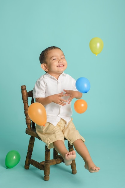 Portret van lachende kleine jongen gekleed in het wit spelen met ballonnen zittend op een houten stoel Blauwe achtergrond