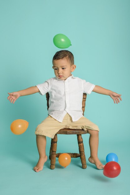 Portret van lachende kleine jongen gekleed in het wit spelen met ballonnen zittend op een houten stoel Blauwe achtergrond