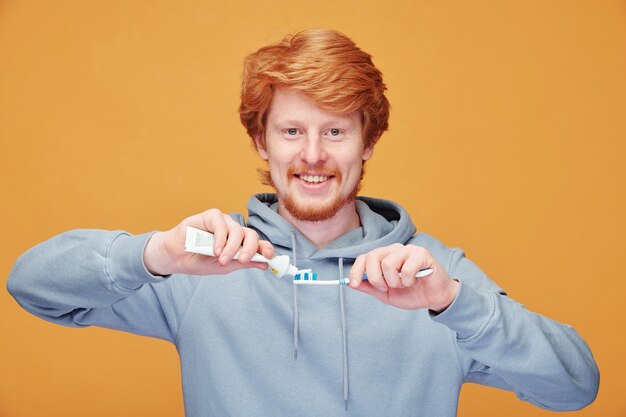 Portret van lachende knappe jonge roodharige man in hoodie tandpasta op tandenborstel toe te passen op oranje