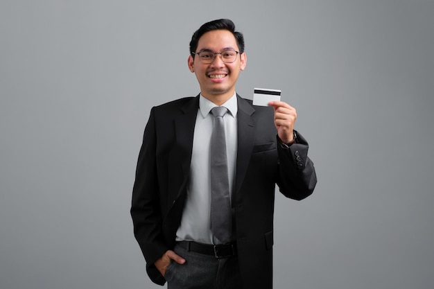 Portret van lachende knappe jonge zakenman in formeel pak met creditcard geïsoleerd op een grijze achtergrond