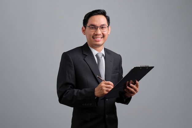 Portret van lachende knappe jonge zakenman in formeel pak met klembord geïsoleerd op een grijze achtergrond
