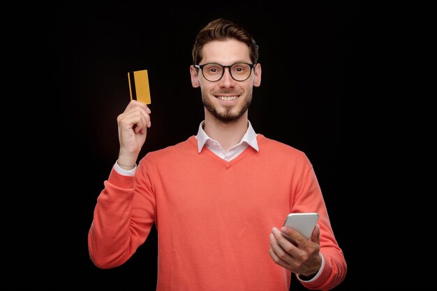 Portret van lachende knappe jongeman in bril met behulp van smartphone-app tijdens het controleren van het saldo van de creditcard