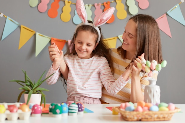 Portret van lachende moeder en dochter met konijnenoren die samen spelen en plezier hebben terwijl ze zich voorbereiden op Pasen tegen een grijze versierde muurfamilie die vrolijk lacht