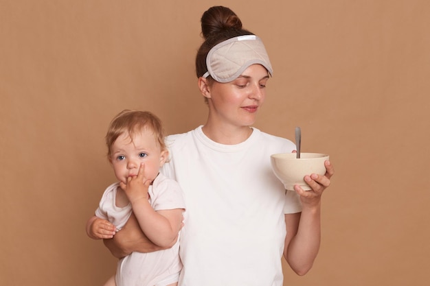 Portret van lachende moeder met baby dochter poseren samen geïsoleerd over bruine achtergrond mama bedrijf bord met puree of pap moet haar kind in de ochtend voeden