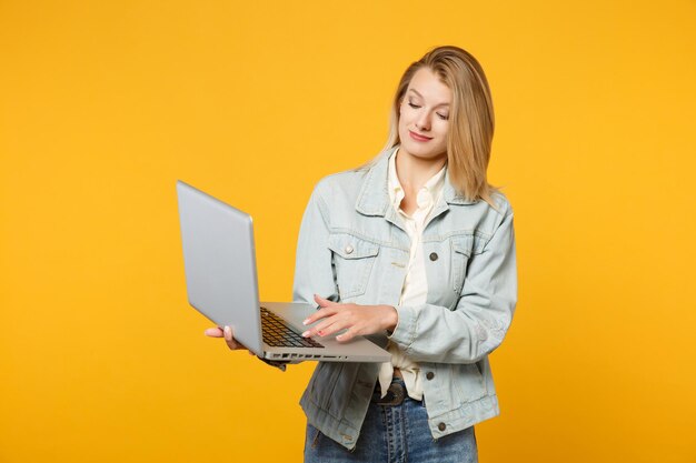 Portret van lachende mooie jonge vrouw in denim casual kleding houden, met behulp van laptop pc-computer geïsoleerd op geel oranje muur achtergrond in studio. Mensen levensstijl concept. Bespotten kopie ruimte.