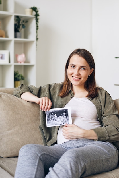 Portret van lachende mooie jonge zwangere vrouw zittend in een ontspannen houding op de bank en poseren met echografie foto van baby