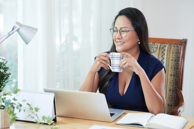Portret van lachende, peinzende, elegante zakenvrouw die een kopje koffie drinkt en wegkijkt wanneer ze geniet van een korte pauze na het werk