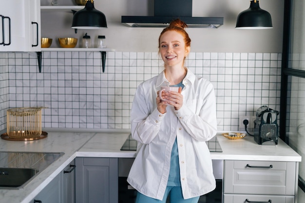 Portret van lachende roodharige vrouw met kopje warme drank in handen op de ochtend in de keuken Aantrekkelijke dame genieten van het drinken van kopje thee thuis Concept van vrijetijdsbesteding roodharige vrouw