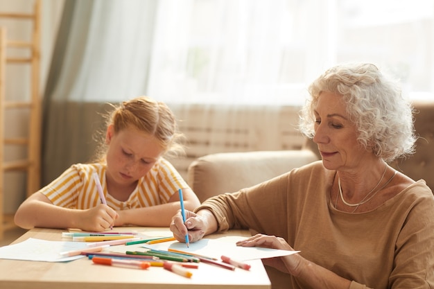 portret van lachende senior vrouw babysitten schattig roodharige meisje en tekening samen zittend bij koffietafel in gezellige woonkamer