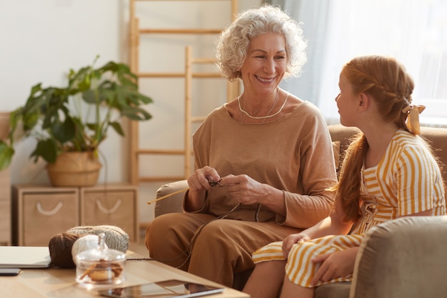 Foto portret van lachende senior vrouw breien met schattig meisje kijkt naar haar in gezellig huis verlicht door zonlicht