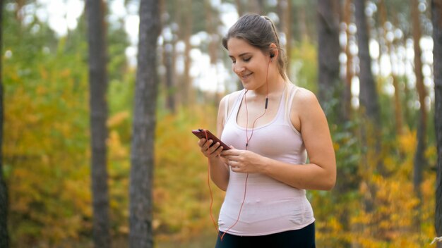 Portret van lachende sportieve vrouw die muzieknummer op smartphone kiest voordat ze jogt of rent in het bos.