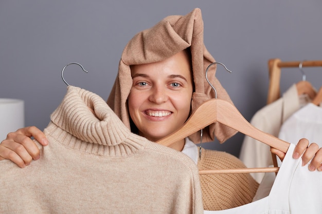 Portret van lachende tevreden jonge volwassen vrouw met kleding op haar hoofd met kleding op twee hangers met stijlvolle kleding die nieuwe outfit kiest en geniet van winkelen