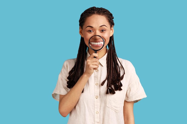 Portret van lachende tevreden vrouw met zwarte dreadlocks met vergrootglas op haar tanden kijken naar camera met gelukkige uitdrukking dragen wit overhemd indoor studio opname geïsoleerd op blauwe achtergrond