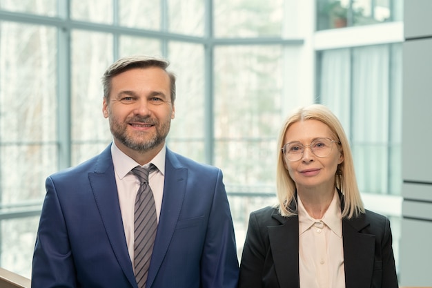 Portret van lachende volwassen zakenmensen in formalwear die tegen een hoog panoramisch raam van een modern kantoor staan