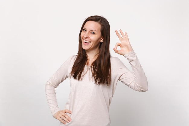 Portret van lachende vrolijke jonge vrouw in lichte kleding op zoek naar camera met OK gebaar geïsoleerd op een witte muur achtergrond in de studio. Mensen oprechte emoties, lifestyle concept. Bespotten kopie ruimte.