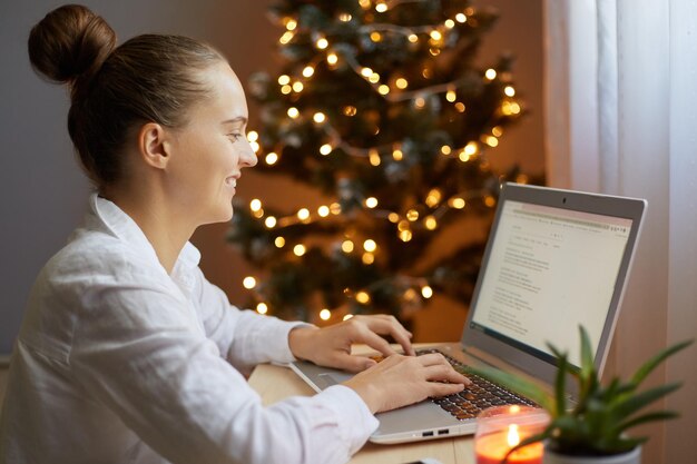 Portret van lachende vrouw die op laptop werkt of online winkelt en nieuwjaarscadeautjes koopt met versierde kerstboom op achtergrond typend op toetsenbord