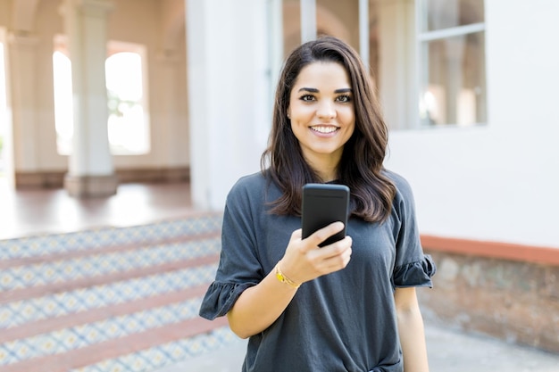 Portret van lachende vrouw die smartphone vasthoudt terwijl ze tegen gebouw staat