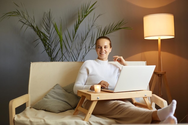 Portret van lachende vrouw, gekleed in witte trui met knotkapsel, zittend op hoest thuis en met laptop kijkend naar film in het weekend kijkend naar de camera