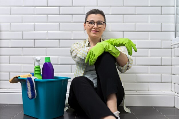 Portret van lachende vrouw in handschoenen zittend op de vloer in de badkamer met emmer wasmiddel. Gelukkige vrouw na het schoonmaken van het huis