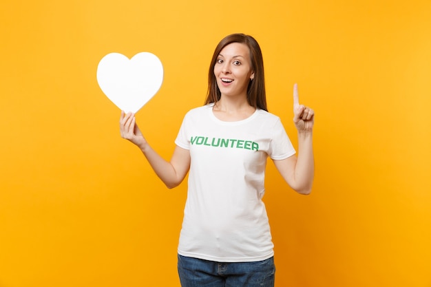 Portret van lachende vrouw in wit t-shirt met geschreven inscriptie groene titel vrijwilliger houdt wit hart geïsoleerd op gele achtergrond. Vrijwillige gratis hulp, liefdadigheidswerkconcept.