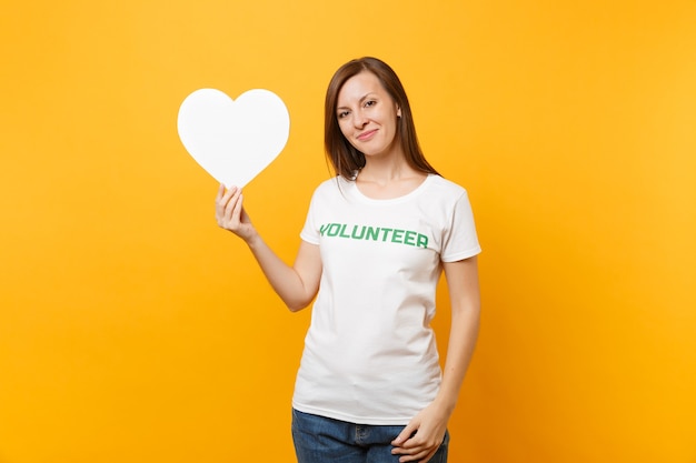 Portret van lachende vrouw in wit t-shirt met geschreven inscriptie groene titel vrijwilliger houdt wit hart geïsoleerd op gele achtergrond. Vrijwillige gratis hulp, liefdadigheidswerkconcept.