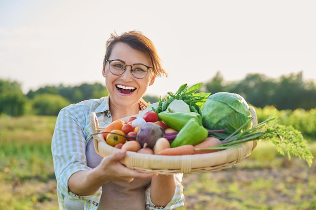 Portret van lachende vrouw van middelbare leeftijd met mand met verschillende verse rauwe groenten en kruiden zomer natuur moestuin achtergrond Oogst van biologische boerderij gezonde voeding voeding tuinieren