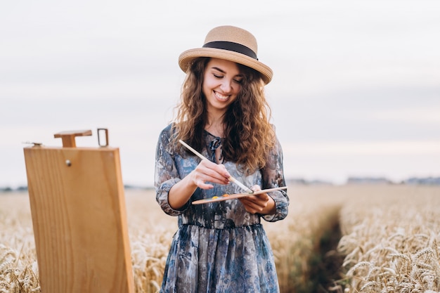 Portret van lachende vrouwelijke artiest met krullend haar in hoed. Meisje tekent een afbeelding van een landschap in een tarweveld. Kopieer ruimte