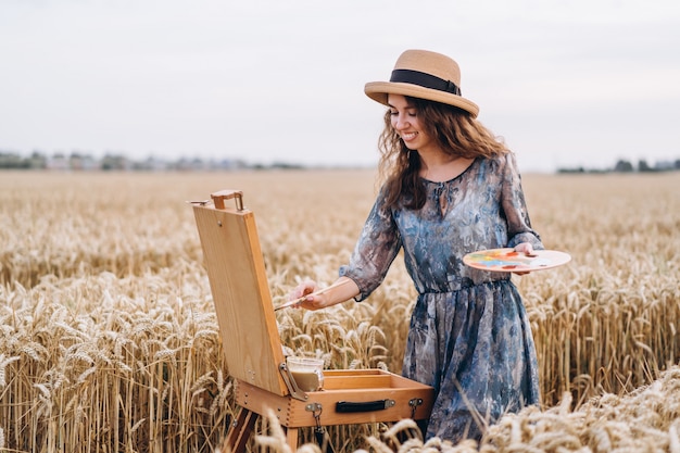 Portret van lachende vrouwelijke artiest met krullend haar in hoed. vrouw tekent een afbeelding van een landschap in een tarweveld. Kopieer ruimte