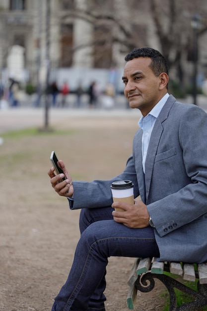 Portret van latino man in jas met een wegwerpbeker koffie zittend op een bankje in een openbaar park