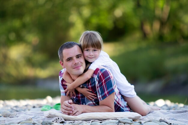 Portret van leuk blond meisje die jonge vader op kiezelsteenrivierbank koesteren op heldere de zomerdag op vage groene bomen. Gelukkig familierelaties, liefde, zorg en perfecte vakantie concept.