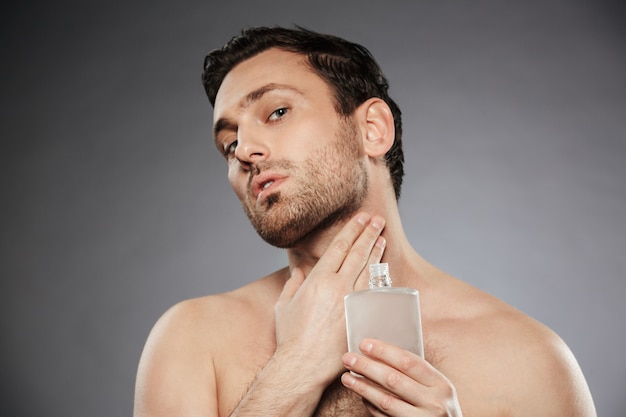 Portret van mannelijke seksuele man parfum aftershave zetten op zijn nek, geïsoleerd over grijze muur