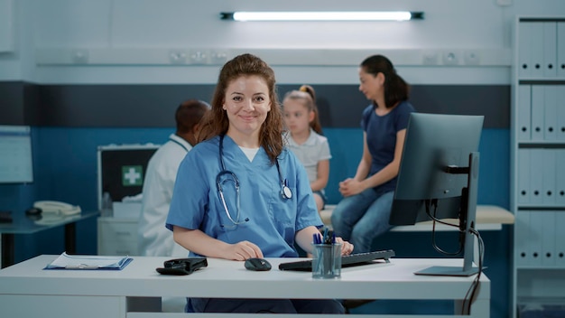 Portret van medisch assistent die computer gebruikt om op afspraak in de kast te werken. vrouw verpleegster met uniform en stethoscoop zittend aan een bureau, bezig met controlebezoek met patiënten.