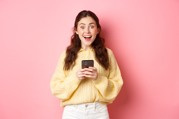 Portret van meisje met smartphone, verbaasd en hijgend opgewonden kijkend, vond online iets interessants, staande tegen een roze muur.