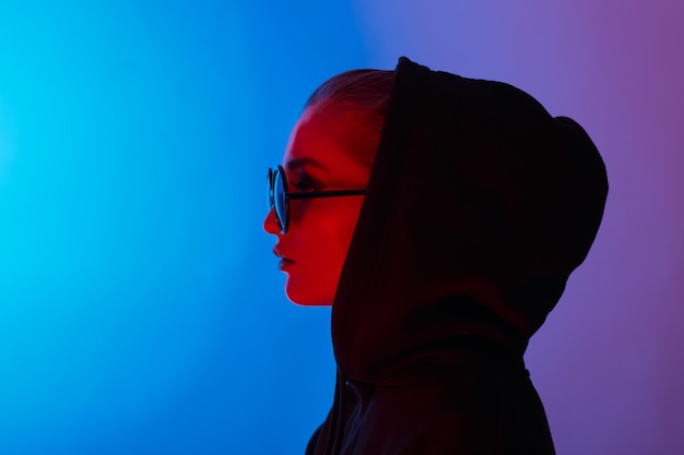 Portret van mode jong meisje in een zwarte trui met een capuchon en ronde zonnebril in rood en blauw neonlicht in de studio.