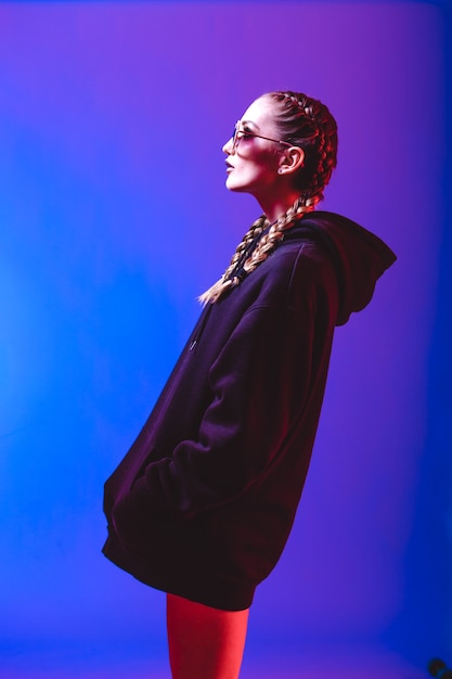 Portret van mode meisje in een zwarte trui met een capuchon en zonnebril ronde vorm in neonlicht in de studio.
